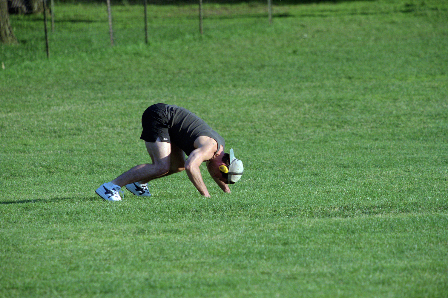 A man working out on all fours in a green field with a hat and headphones on