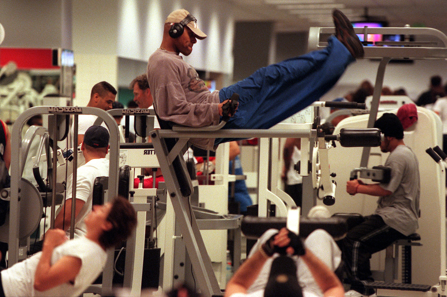A man wearing headphones and a hat on the captain's chair at the gym surrounded by lots of gym-goers