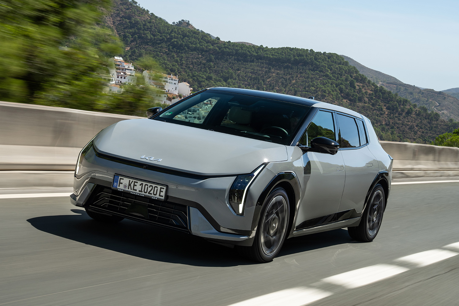 A gray and black car driving down the road with hills and trees behind