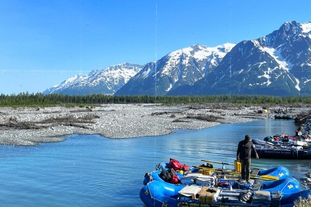 A person stands between two loaded blue rafts numbered 24 and 26 at a sunny river launch site, with a Momentum Alaska raft visible nearby. A braided glacial river winds through a rocky gravel bar, backed by a dense spruce forest and dramatic snow-capped peaks under a clear blue sky.