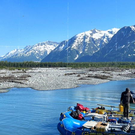 A person stands between two loaded blue rafts numbered 24 and 26 at a sunny river launch site, with a Momentum Alaska raft visible nearby. A braided glacial river winds through a rocky gravel bar, backed by a dense spruce forest and dramatic snow-capped peaks under a clear blue sky.