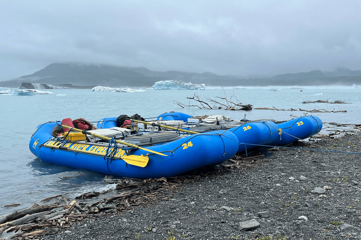 Two blue inflatable rafts numbered 24, 25 and 26, loaded with gear and yellow oars, beached on a rocky shoreline with driftwood. In the background, pale blue icebergs float on a gray-green glacial lake beneath overcast, fog-shrouded mountains.
