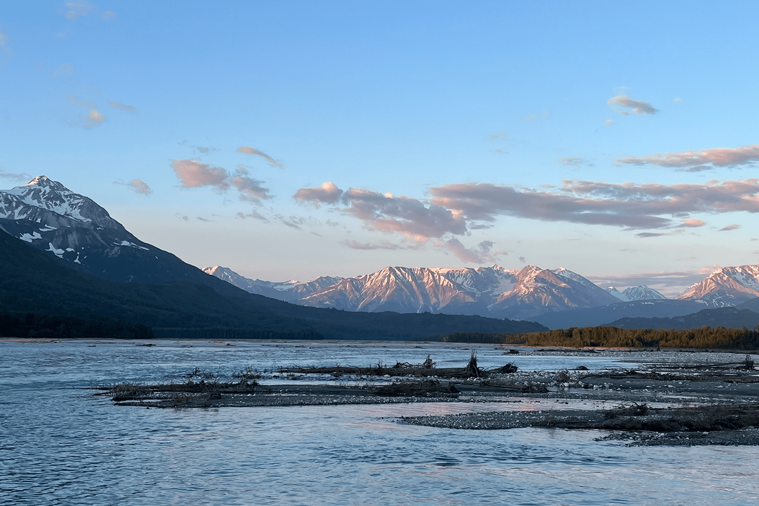 A wide, braided river flows past a driftwood-covered gravel bar at dusk, with golden-lit forest islands in the middle distance and a panoramic range of snow-capped mountains glowing pink and amber under a soft blue sky scattered with wispy clouds.