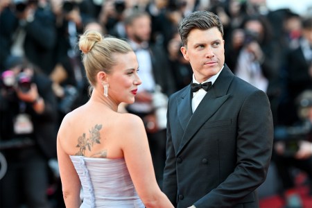 A man in a black tuxedo on a red carpet with a woman in a purple dress with her back turned