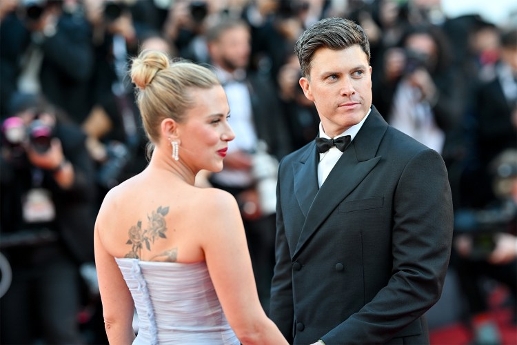 A man in a black tuxedo on a red carpet with a woman in a purple dress with her back turned