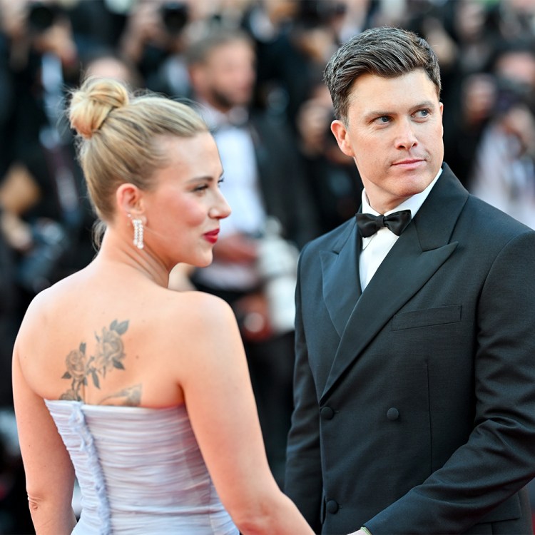 A man in a black tuxedo on a red carpet with a woman in a purple dress with her back turned