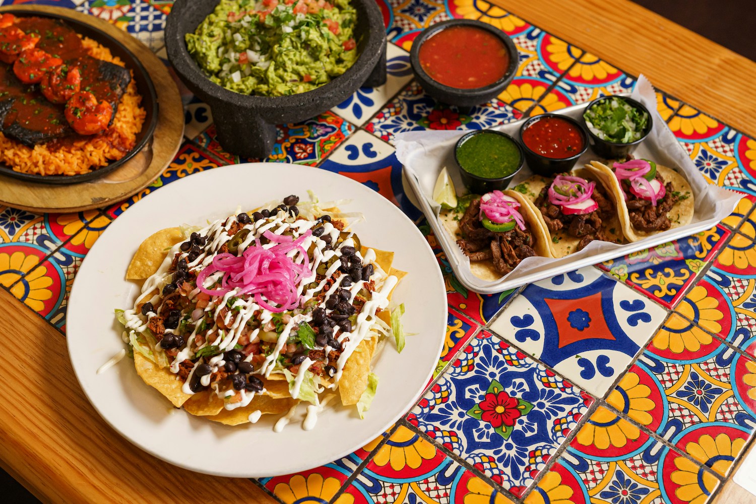 a colorful tile table topped with nachos, tacos, rice, guacamole and salsa