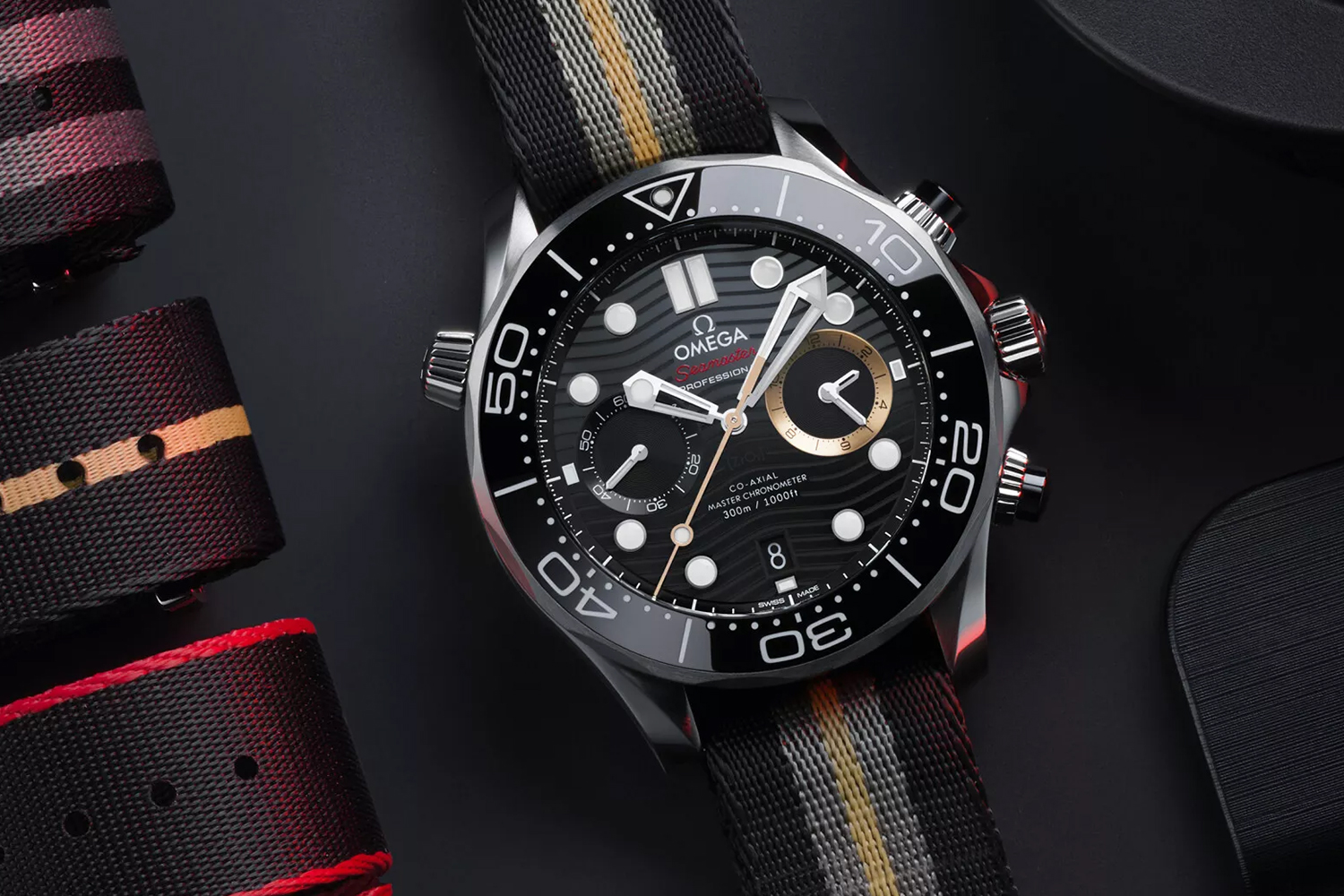 A watch with white hands, a black dial and bezel, and one gold subdial sits on a black table next to folded watch straps