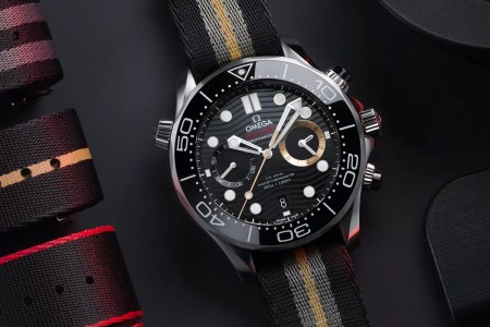 A watch with white hands, a black dial and bezel, and one gold subdial sits on a black table next to folded watch straps