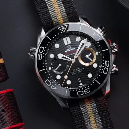 A watch with white hands, a black dial and bezel, and one gold subdial sits on a black table next to folded watch straps