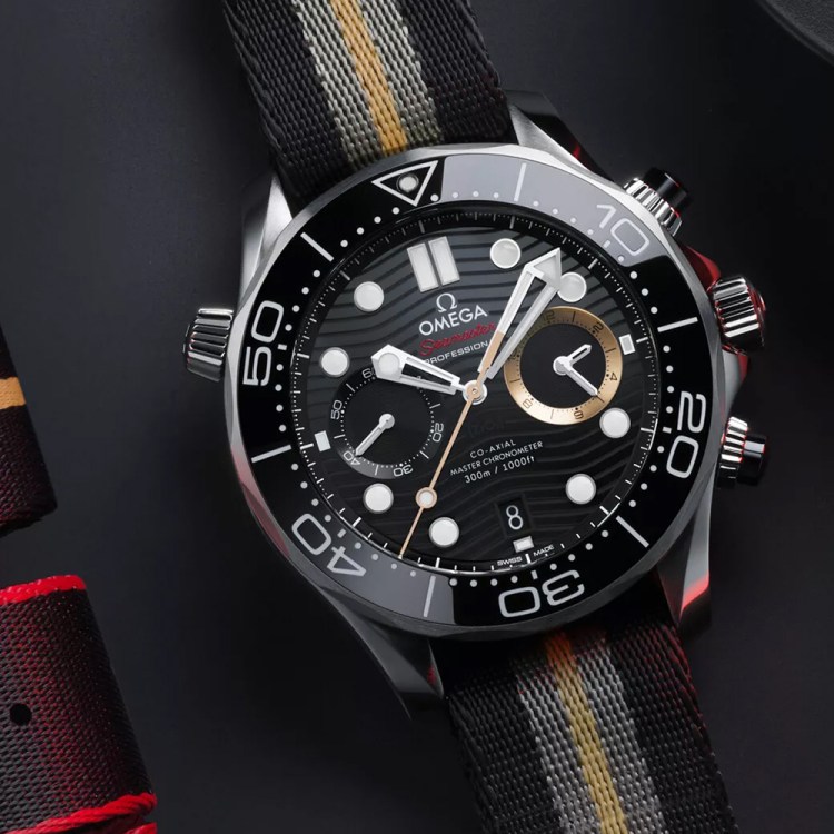 A watch with white hands, a black dial and bezel, and one gold subdial sits on a black table next to folded watch straps