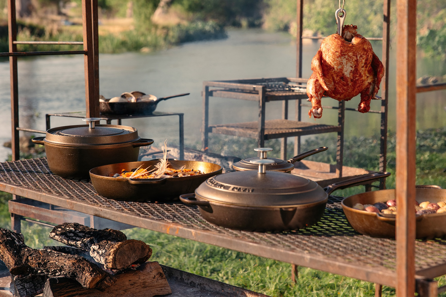 A large, open wood-fired grill with logs underneath a surface holding cast iron cookware