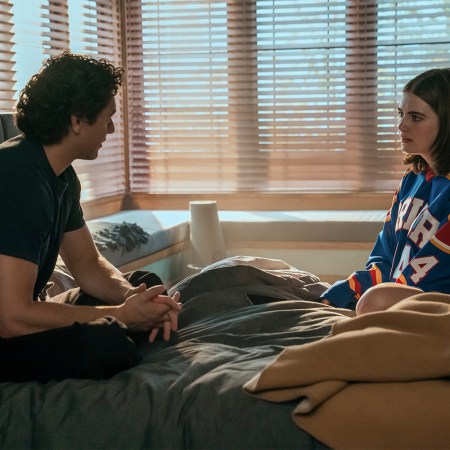 A high school boy in a dark T-shirt sits on a bed with a high school girl in a hockey jersey