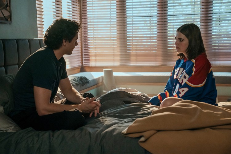 A high school boy in a dark T-shirt sits on a bed with a high school girl in a hockey jersey