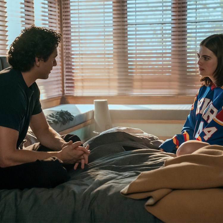 A high school boy in a dark T-shirt sits on a bed with a high school girl in a hockey jersey