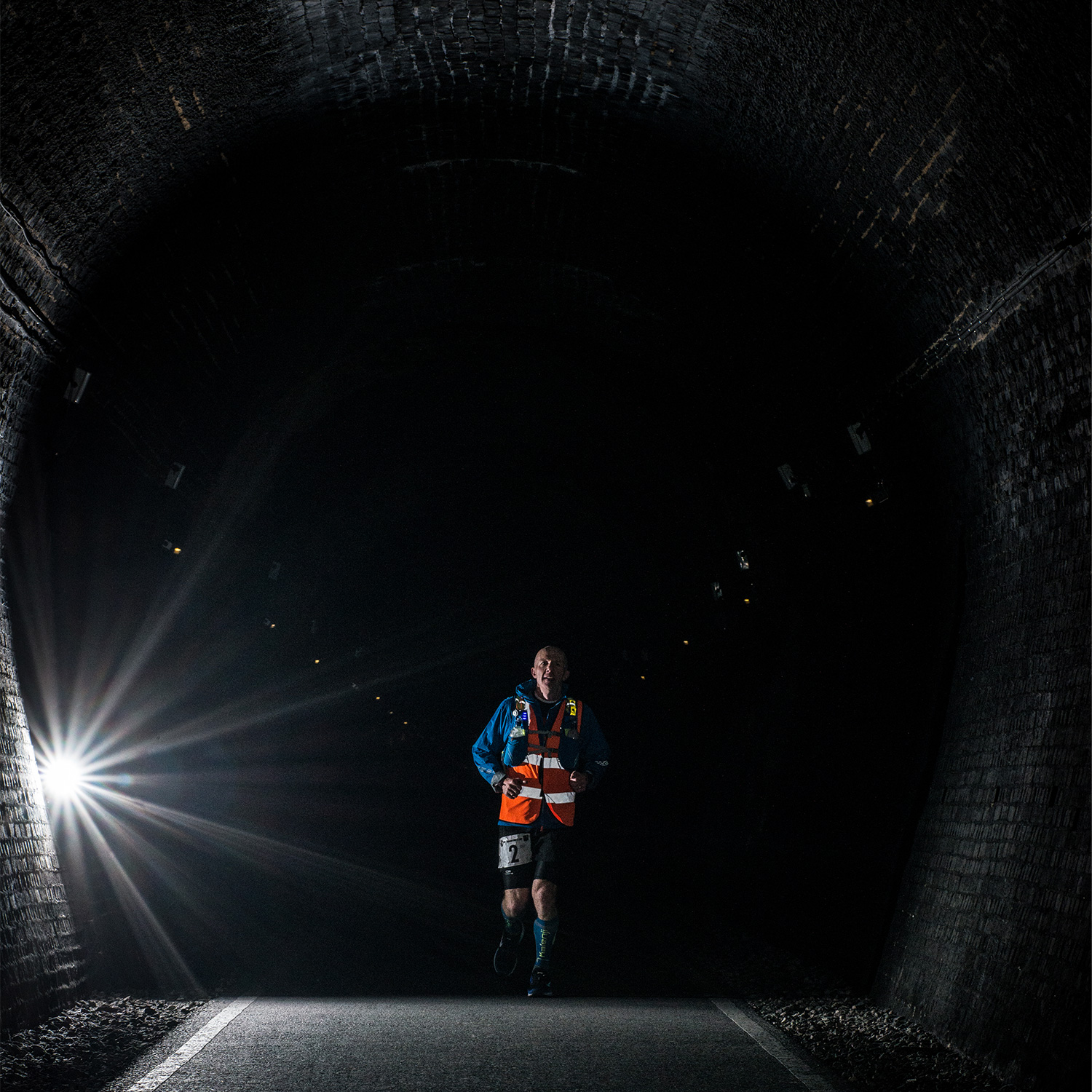 A main in an orange vest running through a dark tunnel with one single light on the left side