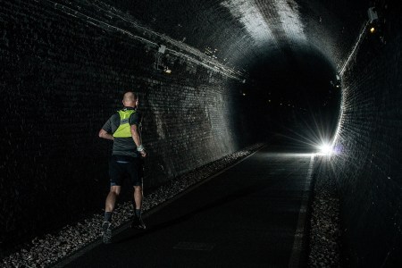 A man running in a high-vis vest through a dark tunnel with a single light shining in the distance