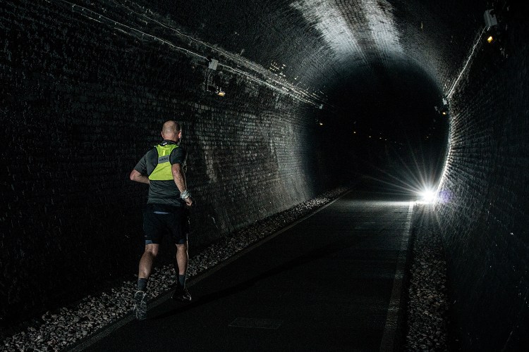 A man running in a high-vis vest through a dark tunnel with a single light shining in the distance