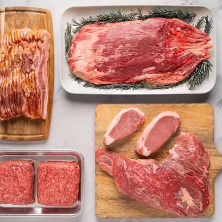 ground beef, pork chops, flank steak and bacon on trays and wooden cutting boards on a white marble surface