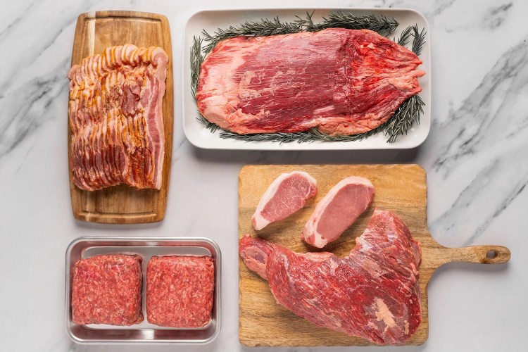 ground beef, pork chops, flank steak and bacon on trays and wooden cutting boards on a white marble surface