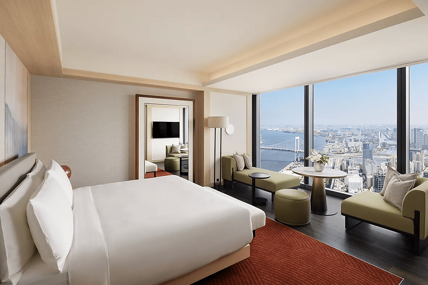 Bright hotel guest room with a king bed in white linens, olive green seating area and dark hardwood floors over a rust-red rug, with floor-to-ceiling windows framing a daytime aerial view of Tokyo Bay and Rainbow Bridge.