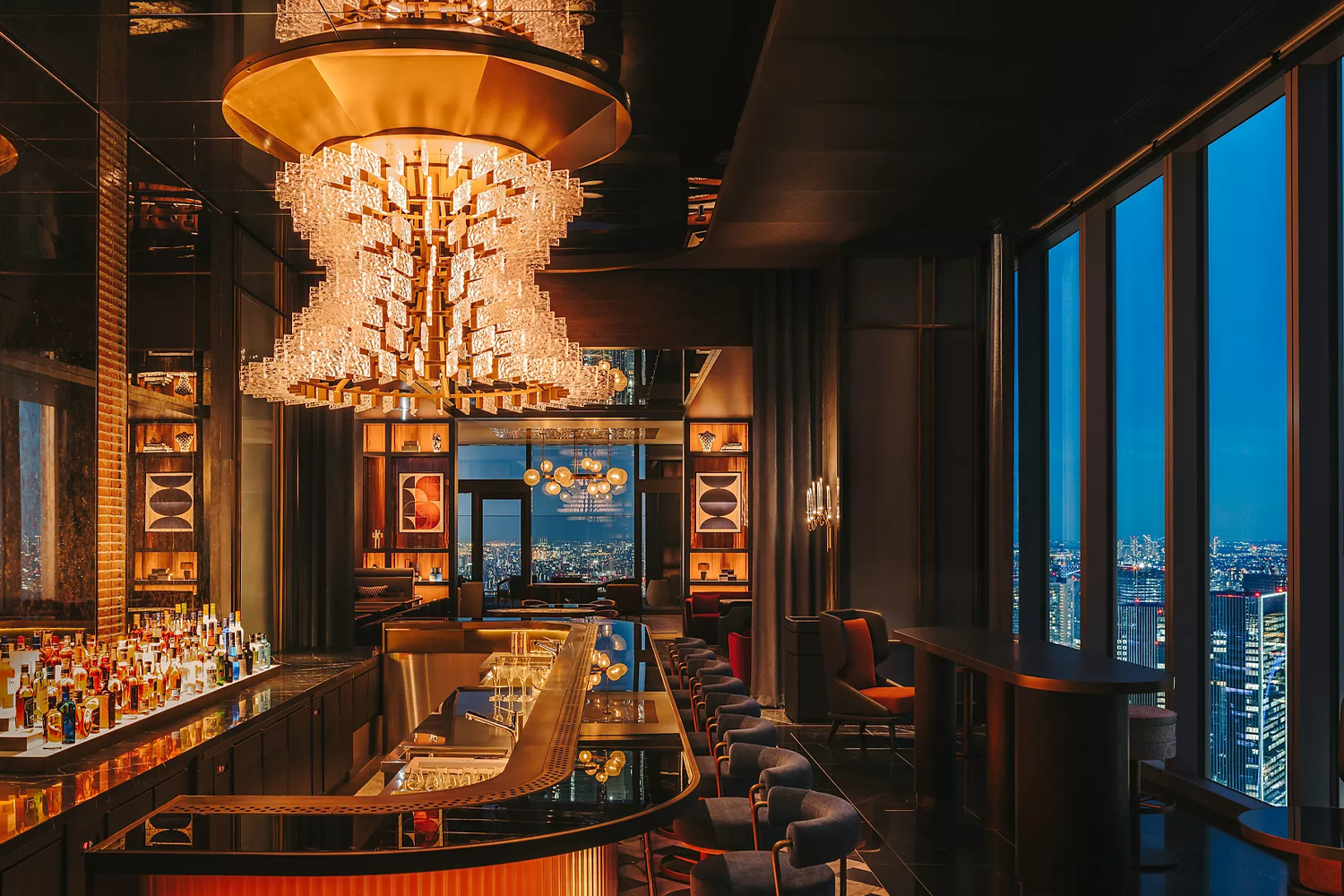 The warmly lit bar at Driftwood Bar & Restaurant in the Fairmont Tokyo, with a cascading crystal chandelier overhead, a curved black marble bar stocked with spirits and floor-to-ceiling windows overlooking the glittering Tokyo skyline at dusk.