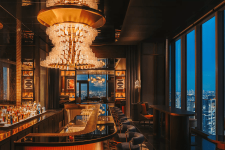 The warmly lit bar at Driftwood Bar & Restaurant in the Fairmont Tokyo, with a cascading crystal chandelier overhead, a curved black marble bar stocked with spirits and floor-to-ceiling windows overlooking the glittering Tokyo skyline at dusk.