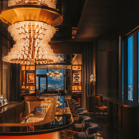 The warmly lit bar at Driftwood Bar & Restaurant in the Fairmont Tokyo, with a cascading crystal chandelier overhead, a curved black marble bar stocked with spirits and floor-to-ceiling windows overlooking the glittering Tokyo skyline at dusk.