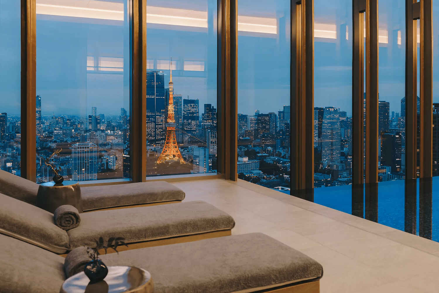 Interior of a hotel spa relaxation room at dusk, with two padded lounge beds in the foreground and floor-to-ceiling bronze-framed windows overlooking the illuminated Tokyo Tower and city skyline at blue hour.