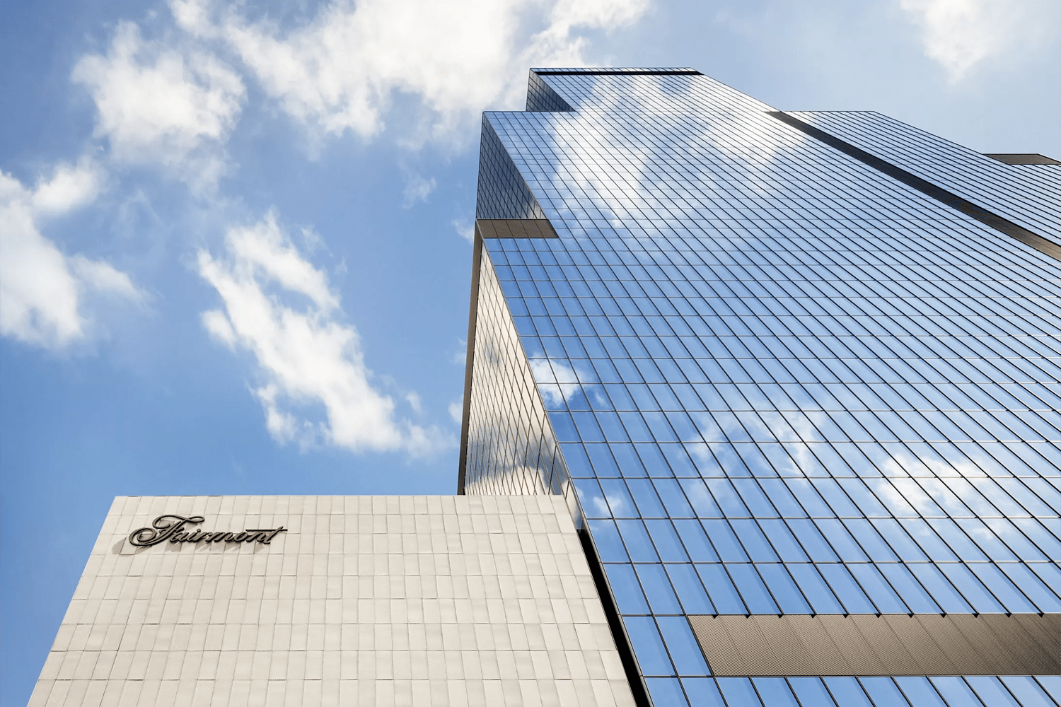 Low-angle exterior shot of the Fairmont Tokyo, showing a soaring glass-curtain-wall tower reflecting clouds and blue sky, alongside a lower cream-stone podium bearing the Fairmont script logo in black.