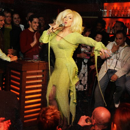 A drag performer in a sparkling yellow-green gown and platinum blonde wig sings into a microphone on the floor of a packed bar, surrounded by a cheering crowd.