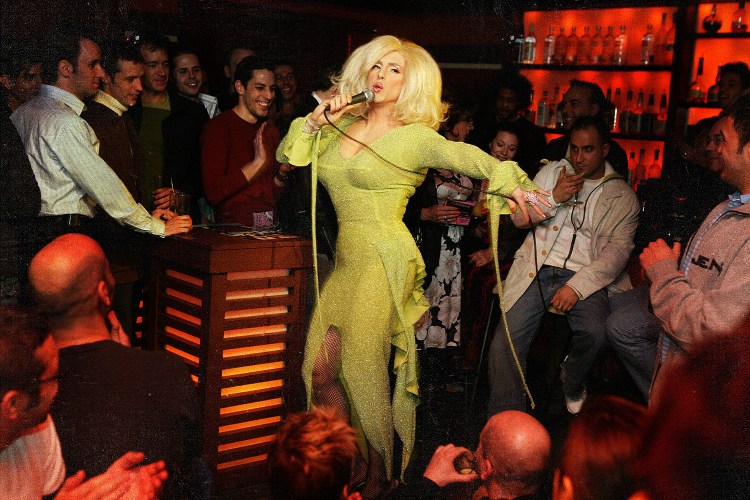 A drag performer in a sparkling yellow-green gown and platinum blonde wig sings into a microphone on the floor of a packed bar, surrounded by a cheering crowd.