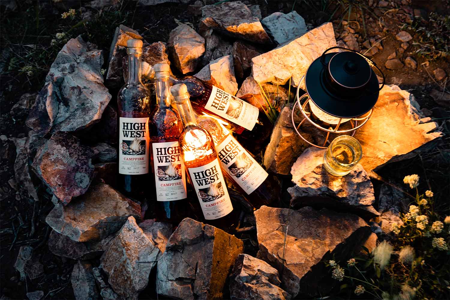 Several bottles of High West Campfire whiskey on an unlit, makeshift fire pit with a lantern and a glass of whiskey