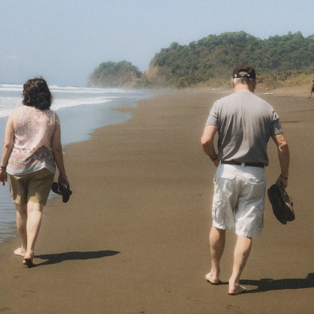 An older couple walks barefoot along a dark-sand beach at the water's edge, shoes in hand, green headlands rising behind them