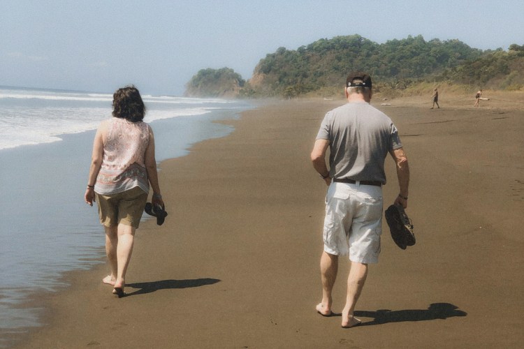 An older couple walks barefoot along a dark-sand beach at the water's edge, shoes in hand, green headlands rising behind them