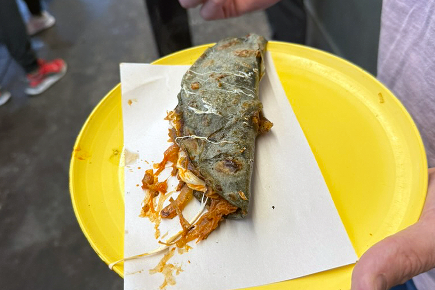 A griddle-pressed blue corn quesadilla, its tortilla darkened and streaked with melted cheese, sits on a sheet of white paper atop a bright yellow plastic plate, stuffed with caramelized onions spilling out the sides — held by hand at a busy street market.
