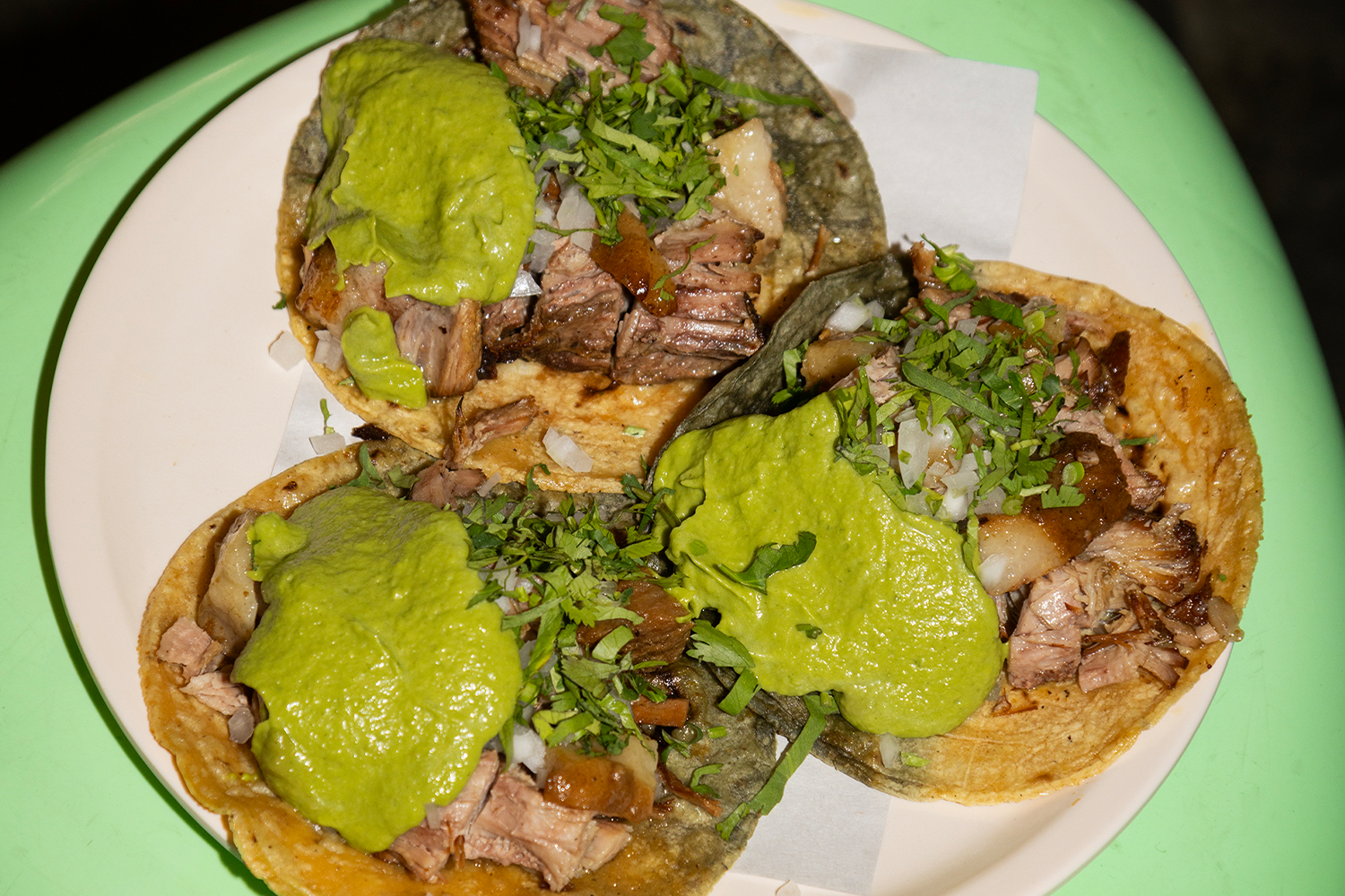 Four tacos on a white paper-lined plate — a mix of blue and yellow corn tortillas piled with braised meat, diced onion, and chopped cilantro, each topped with a generous dollop of vivid green avocado salsa, shot from above against a bright mint-green table.