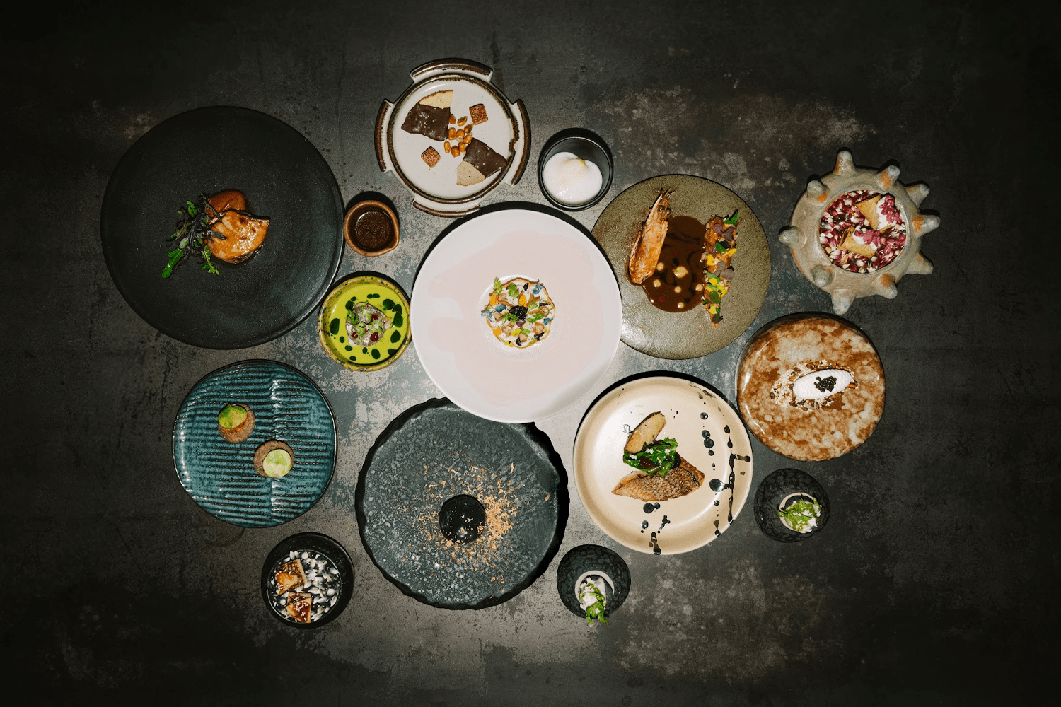 An overhead spread of an entire tasting menu at Maizajo, with a dozen dishes fanned across a dark concrete surface — each course plated on a different artisanal vessel, from a jagged volcanic stone slab to a spiky ceramic bowl, showcasing composed bites of seafood, chocolate, caviar, and vegetables in miniature.