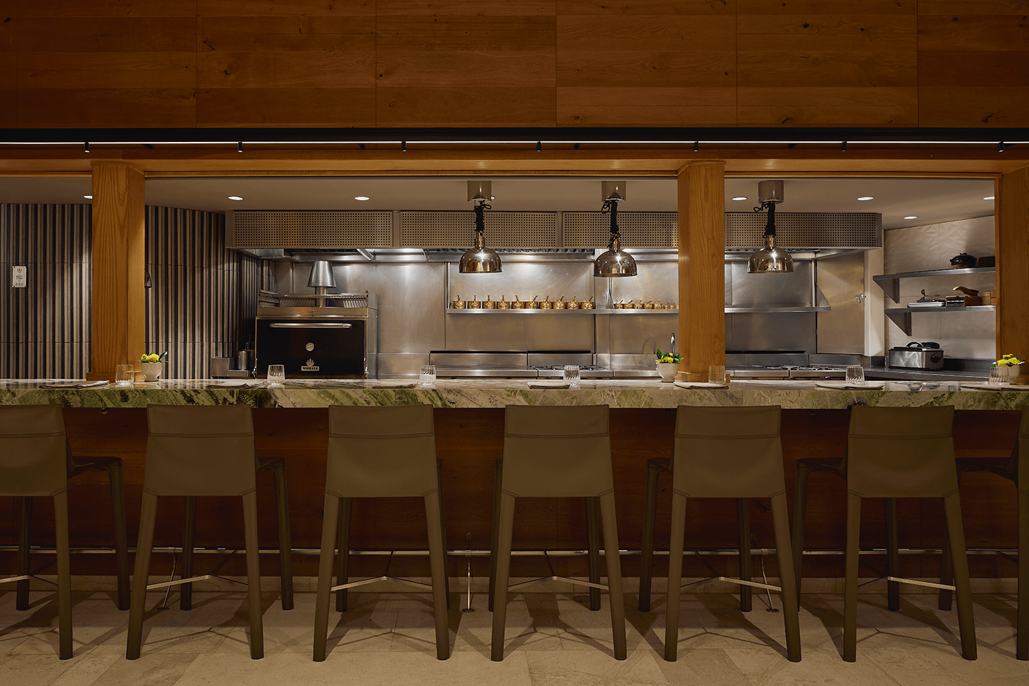 The chef's counter at Quintonil, lined with olive-green leather bar stools and topped with a veined green marble surface, looks directly into the restaurant's open kitchen, where copper heat lamps hang above stainless steel prep stations and a black oven glows against warm wood-paneled walls.