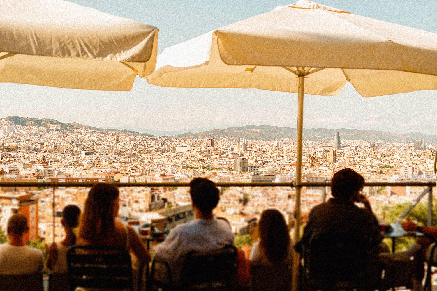 Travelers seated at an outdoor terrace café beneath white umbrellas, looking out over a sweeping panoramic view of Barcelona's skyline and rooftops, with mountains in the distance.