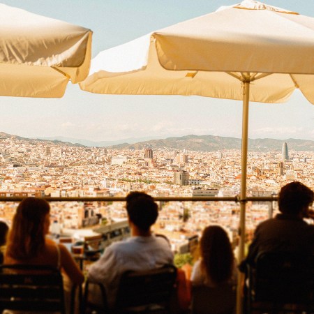 Travelers seated at an outdoor terrace café beneath white umbrellas, looking out over a sweeping panoramic view of Barcelona's skyline and rooftops, with mountains in the distance.