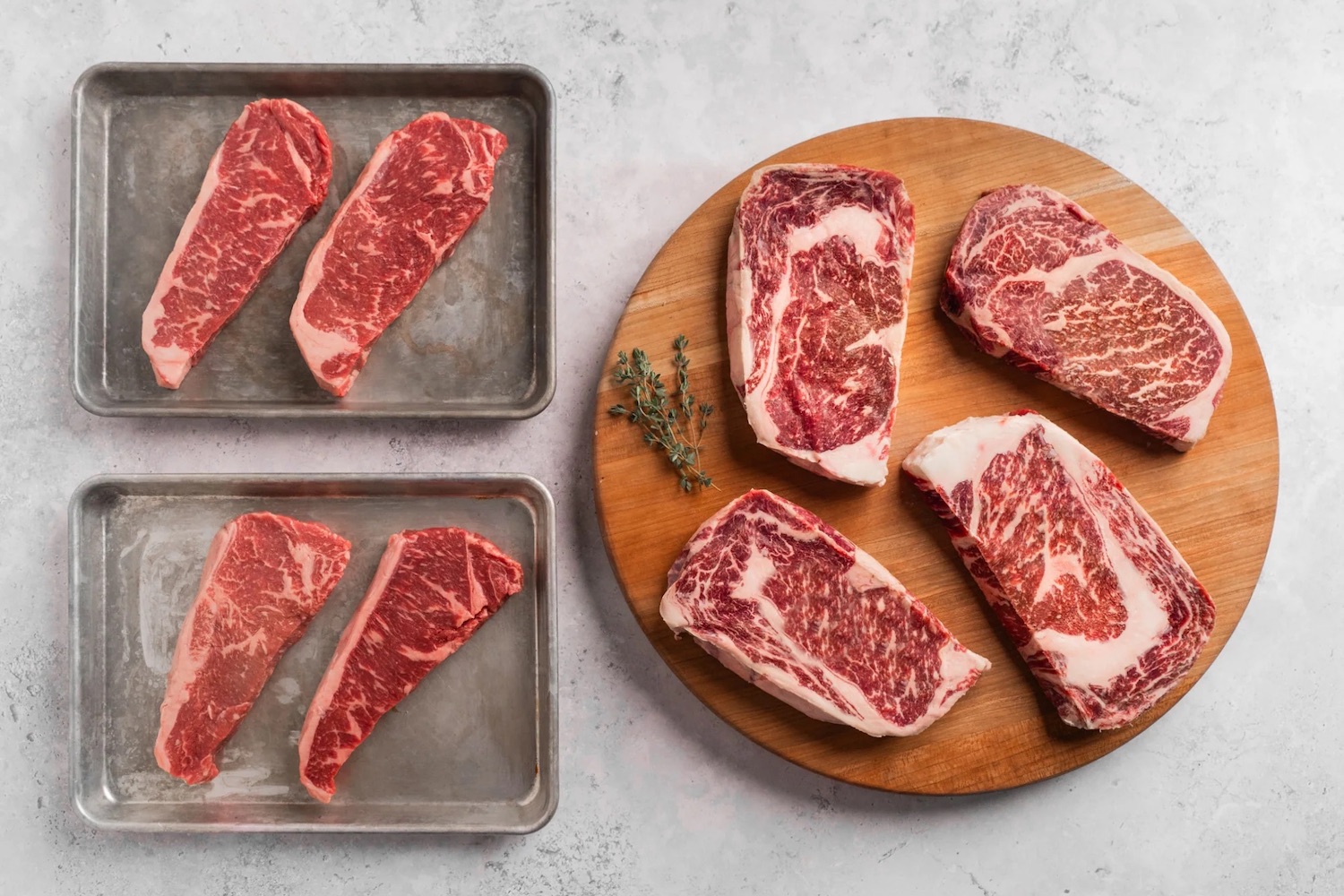two trays of steaks and a wooden cutting board with more steaks on a white marble surface