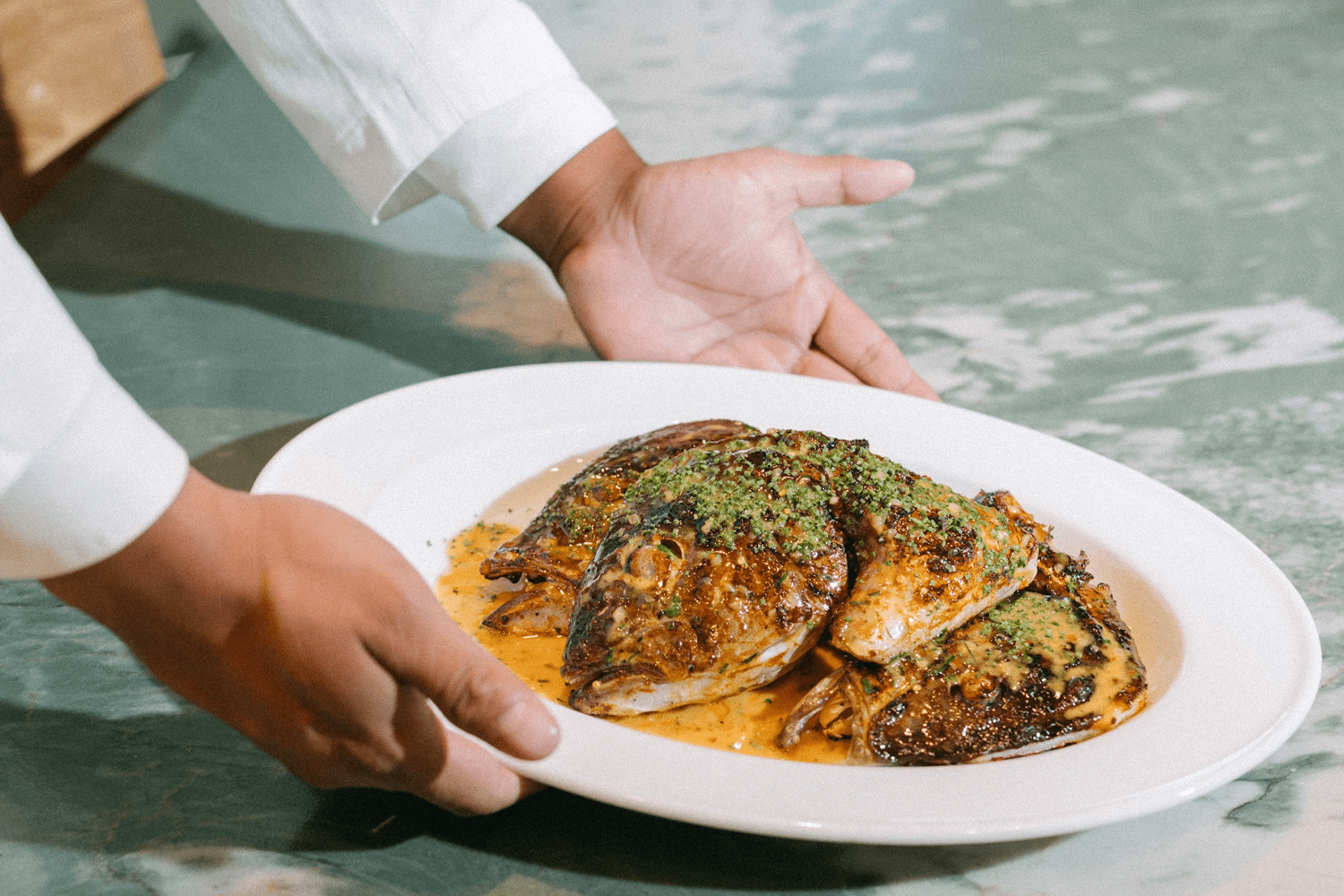 A server in a white chef's coat presents a whole roasted fish on a wide shallow bowl, its skin charred and glistening, topped with a bright green herb crust and pooled in a golden butter sauce, set against a sea-green marble surface.