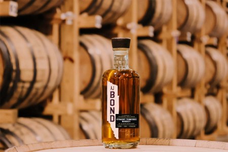 A bottle of AJ Bond whiskey in front of rows of barrels in a rickhouse