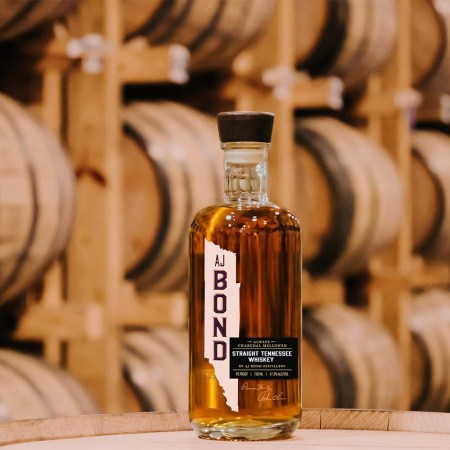 A bottle of AJ Bond whiskey in front of rows of barrels in a rickhouse