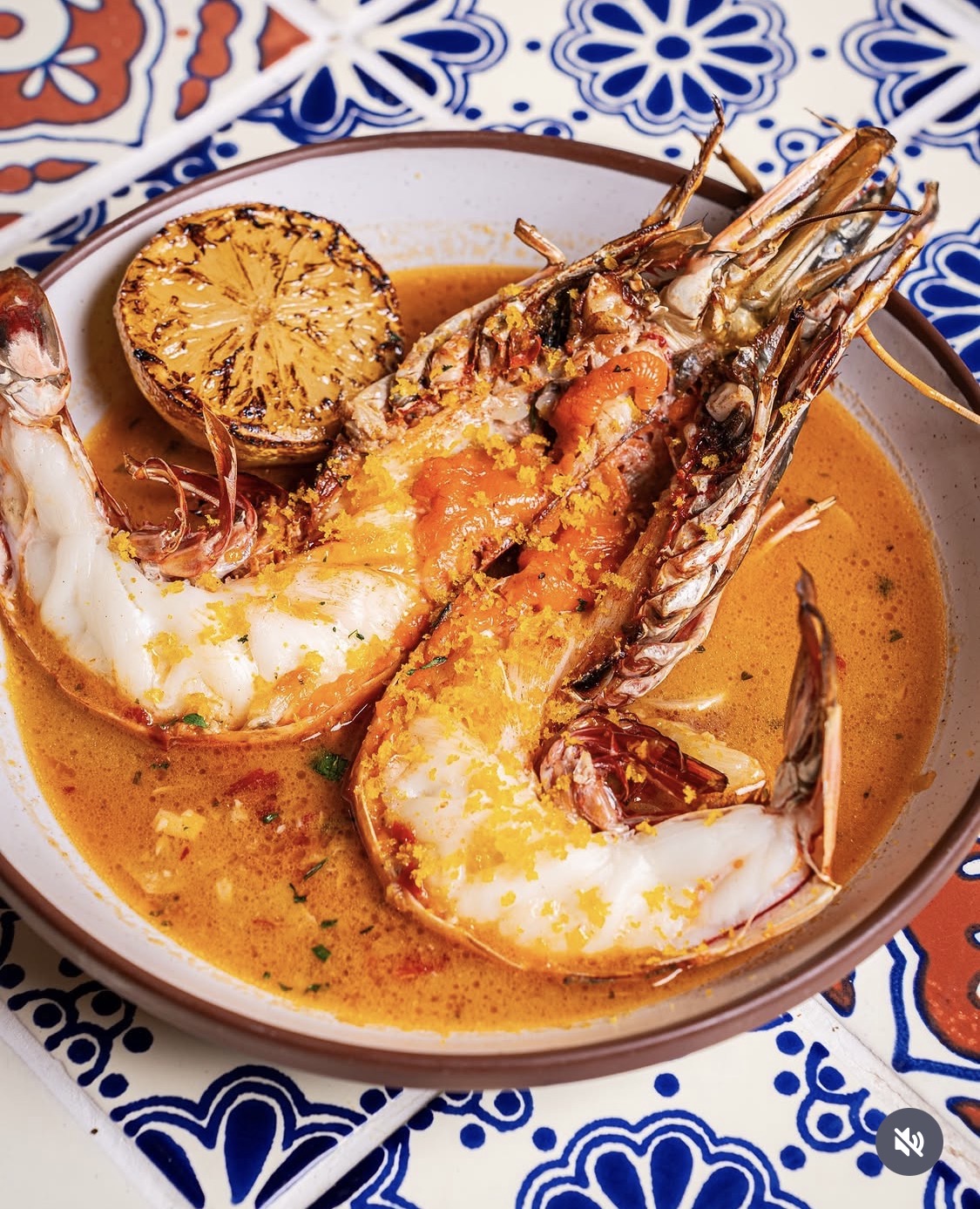 a large split grilled prawn next to a grilled lemon in a red broth on top of a blue and white tiled table