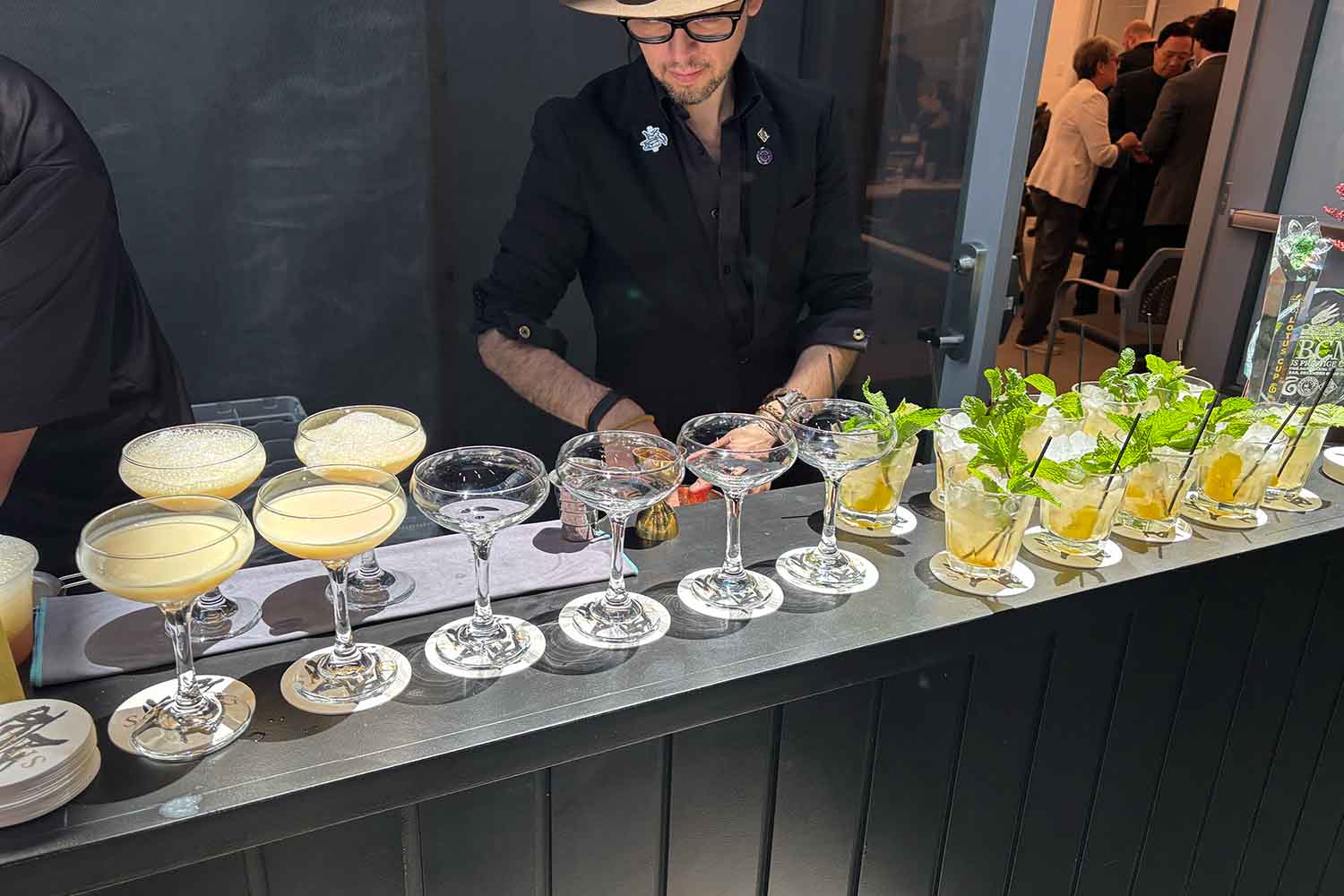 A bartender in a hat serving two types of cocktails on a bar top