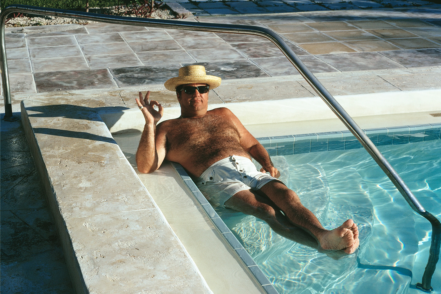 A dad in a straw hat with sunglasses and white swim trunks sitting in the corner of a pool making the OK sign