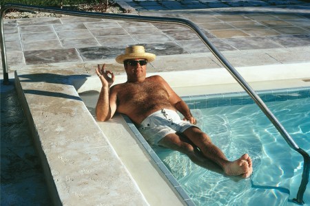 A dad in a straw hat with sunglasses and white swim trunks sitting in the corner of a pool making the OK sign
