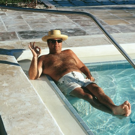 A dad in a straw hat with sunglasses and white swim trunks sitting in the corner of a pool making the OK sign